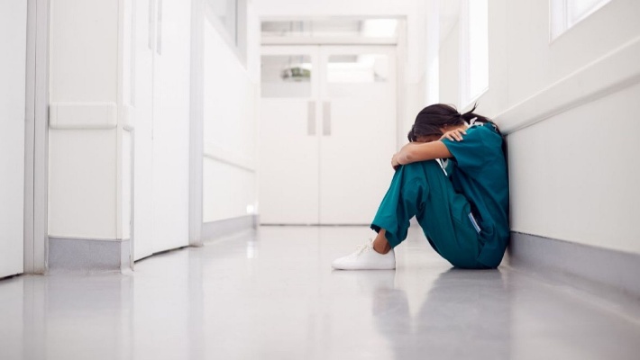 Una sanitaria con pijama de color verde está sentada en el suelo con su cabeza entre suss brazos, que están apoyados sobre las rodillas